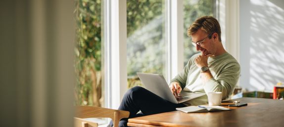 Junger Mann sitzt mit Laptop am Esstisch in der Sonne